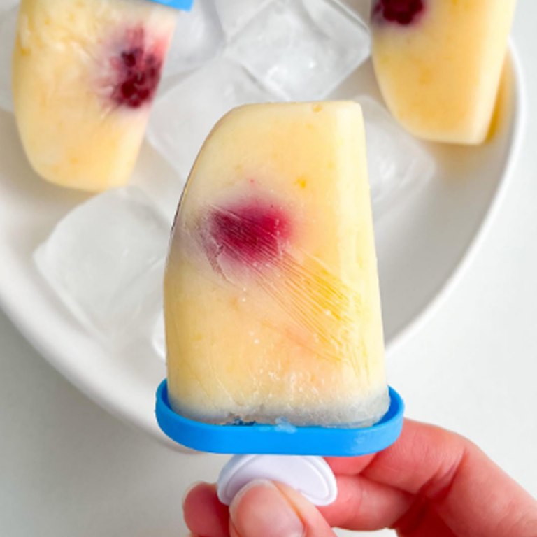 Receita de picolé saudável com apenas dois ingredientes, apresentados em moldes azuis com o pegador em formato de folha, sobre um prato branco em formato de coração com gelo. O picolé tem coloração clara e um detalhe roxo no centro, como uma fruta.