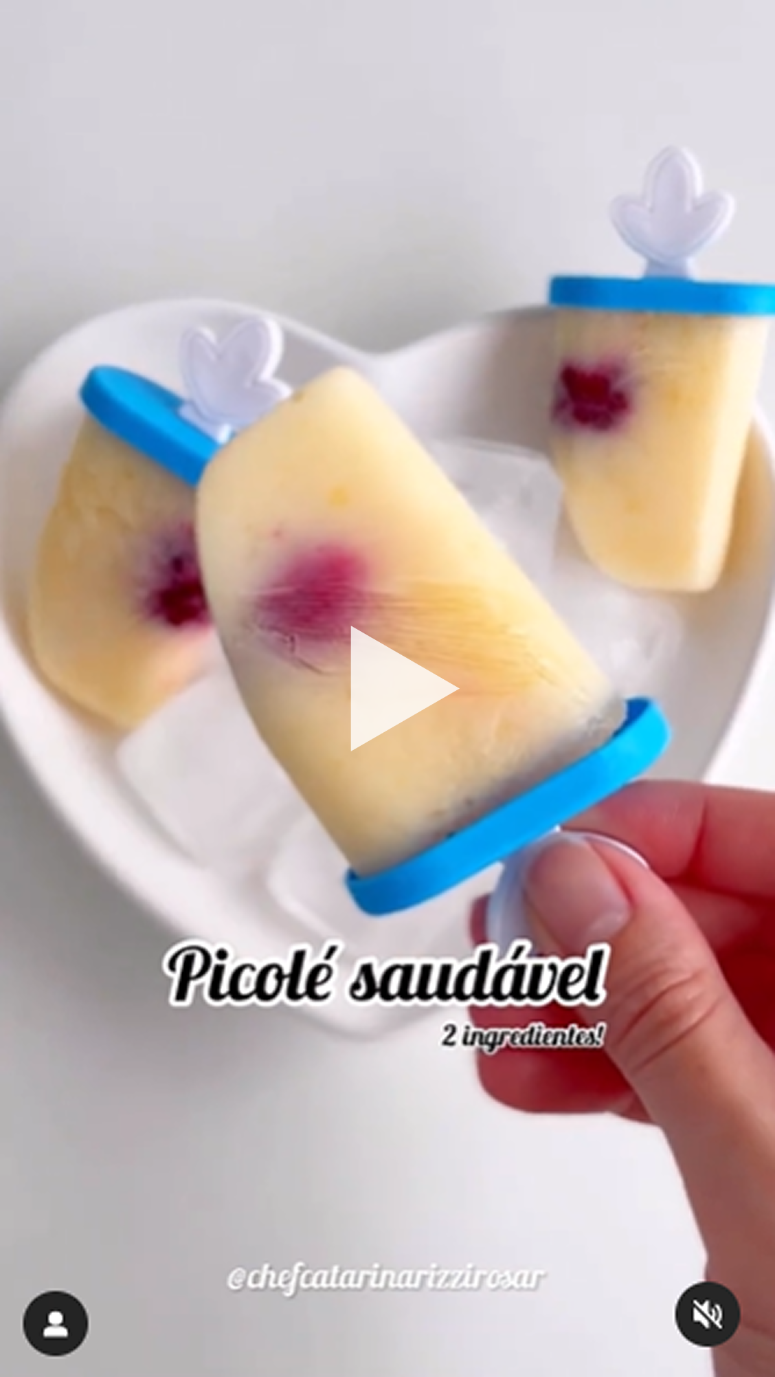 Receita de picolé saudável com apenas dois ingredientes, apresentados em moldes azuis com o pegador em formato de folha, sobre um prato branco em formato de coração com gelo. O picolé tem coloração clara e um detalhe roxo no centro, como uma fruta.