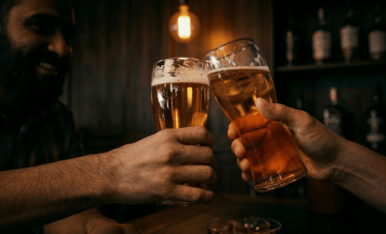 Homens brindando com dois copos de cerveja em um bar com iluminação quente e ambiente aconchegante, enquanto garrafas aparecem desfocadas ao fundo.