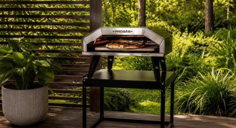 Forno refratário de pizza em inox sobre suporte metálico preto, com pizza assando em seu interior. O equipamento está em uma área externa com deck de madeira, plantas e fundo verde iluminado pela luz natural.